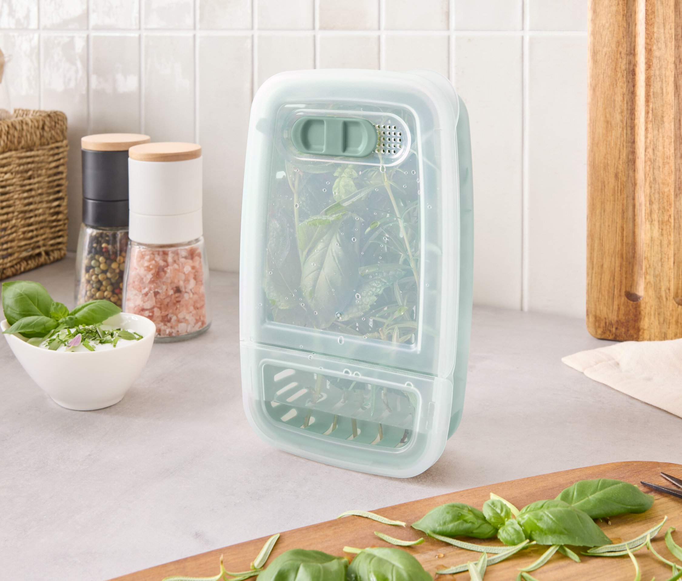 Egy gyógynövénytároló edény gyógynövényekkel a belsejében áll a pulton, hozzávalók és egy vágódeszka mellett.