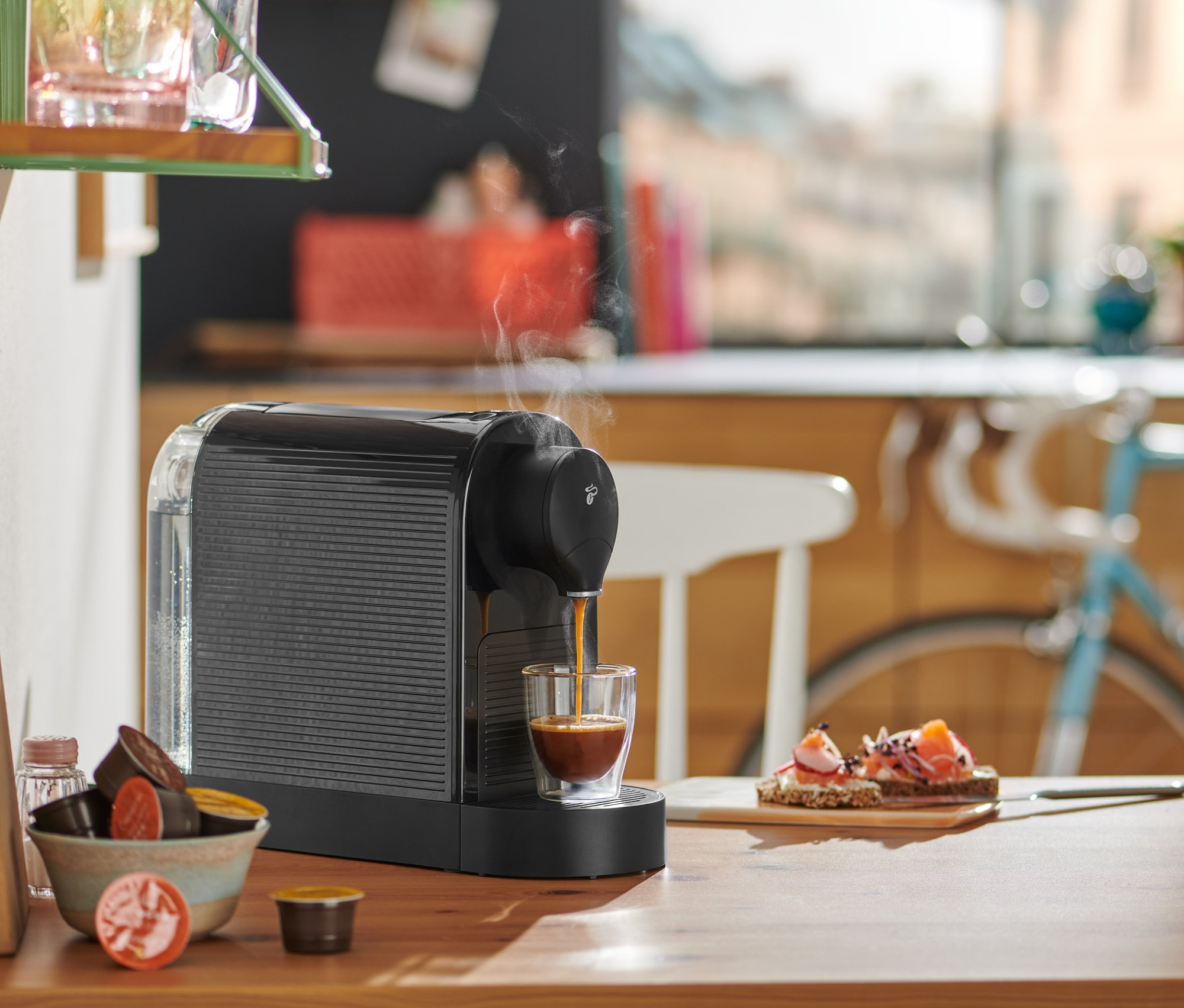 Fekete Cafissimo pure plus kávéfőző eszpresszót készít egy pohárba egy fa asztalon.