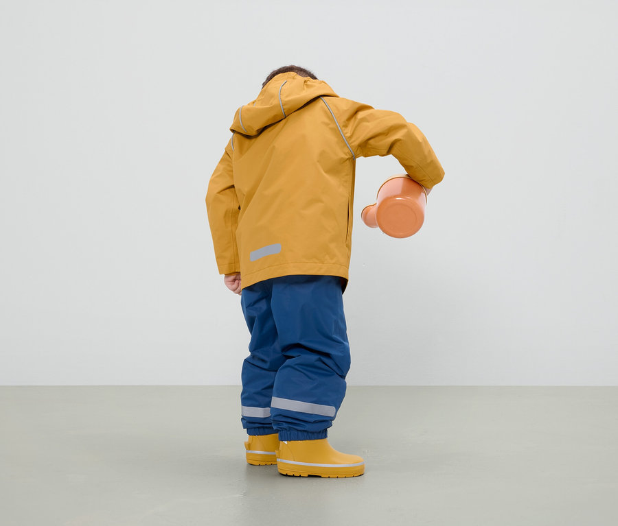 Egy mustársárga kabátot, kék nadrágot és sárga gumicsizmát viselő fiú egy narancssárga locsolókannát tart.