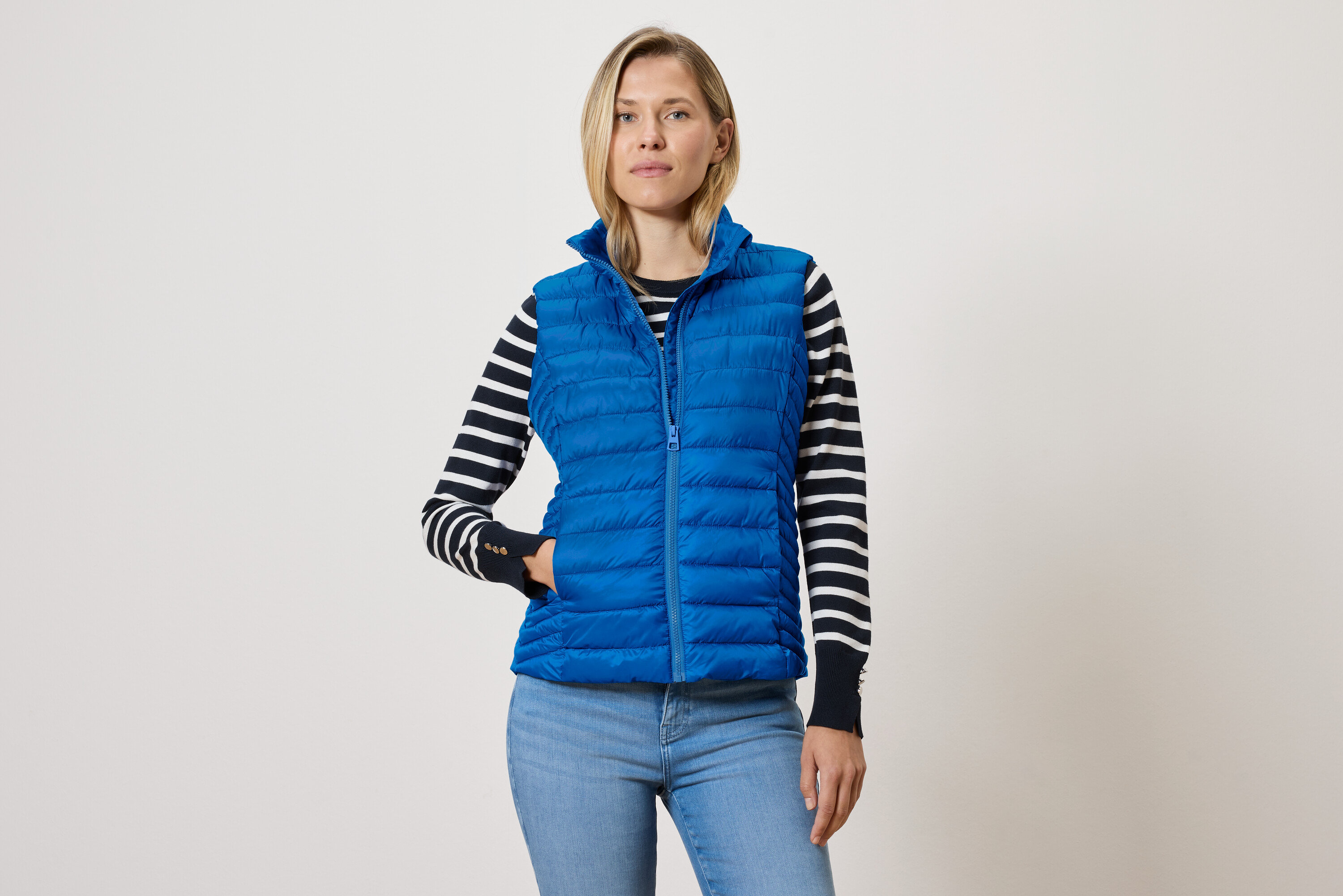 Nő kék, cipzáras, steppelt mellényt, fekete-fehér csíkos pulóvert és kék farmert visel.