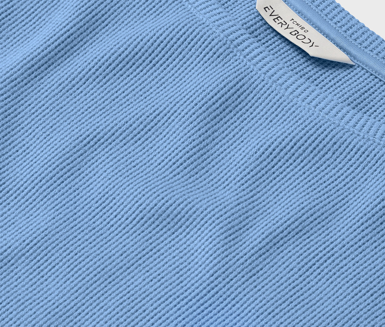 Világoskék, bordázott anyag részlete, fehér címkével, rajta az EVERYBODY felirat.