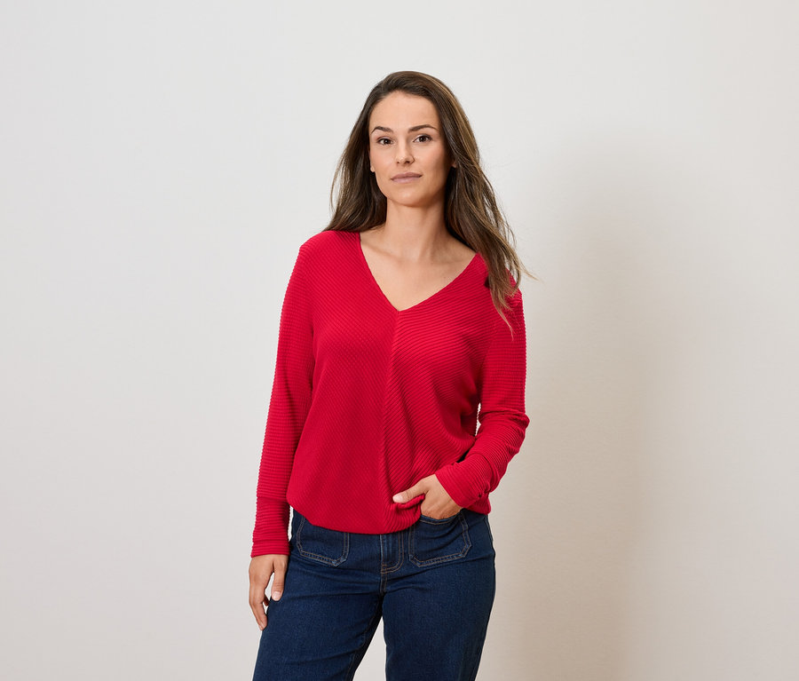 Barna hajú nő pózol piros, hosszú ujjú pulóverben és kék farmerban.