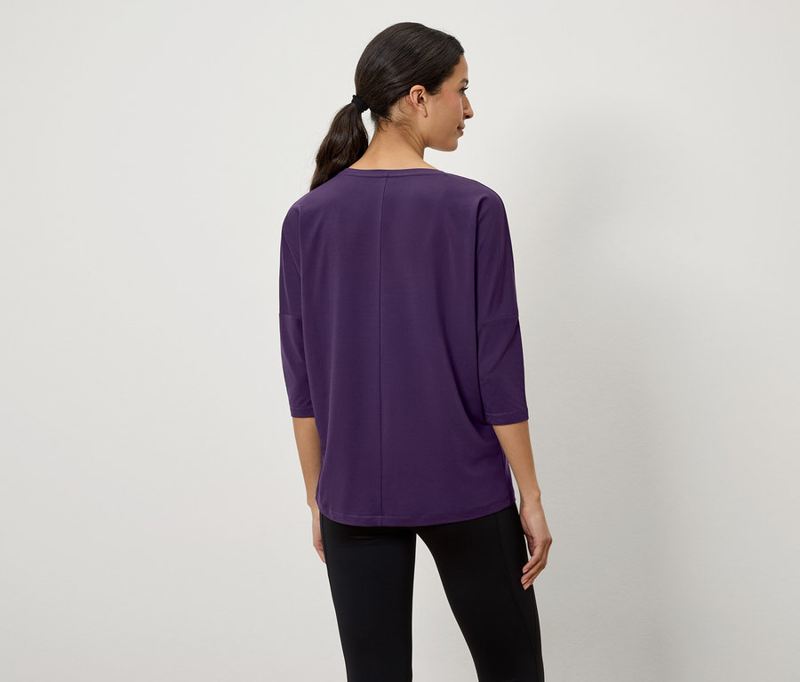 Hátulról látható, sötét hajú, copfba kötött nő lila, háromnegyedes ujjú pólót és fekete leggingset visel.