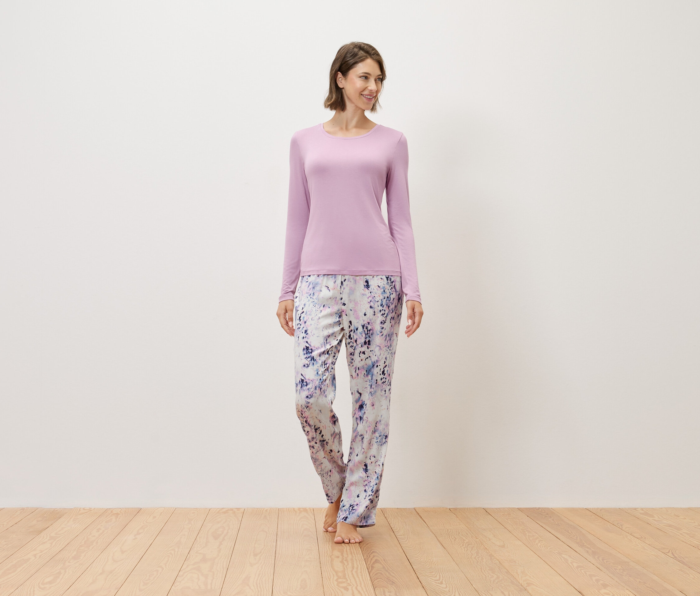 Nő áll mezítláb egy fapadlón, lila pulóvert és mintás nadrágot visel.