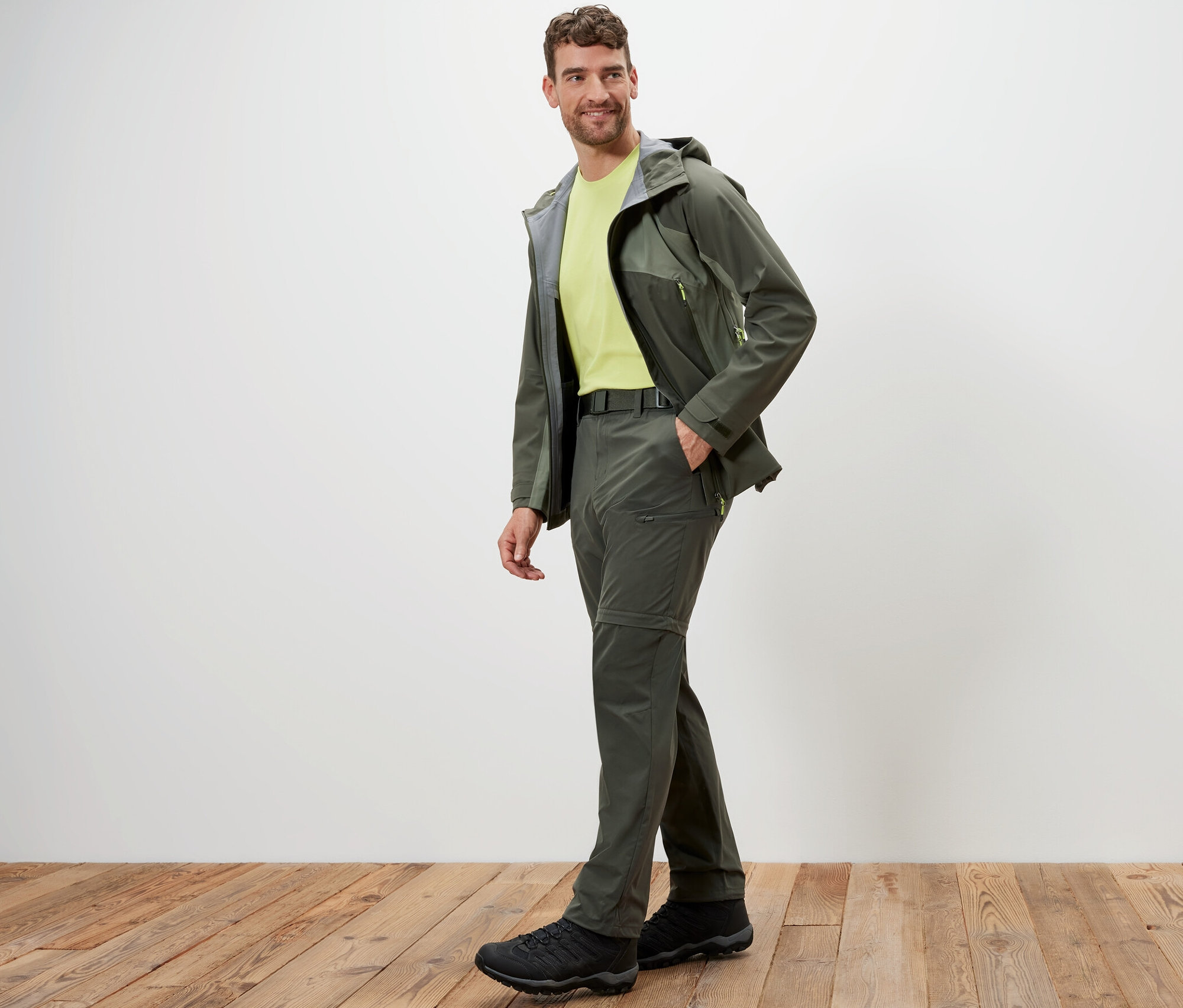 Férfi pózol khaki színű 2 az 1-ben funkcionális nadrágban és kabátban, fapadlón.