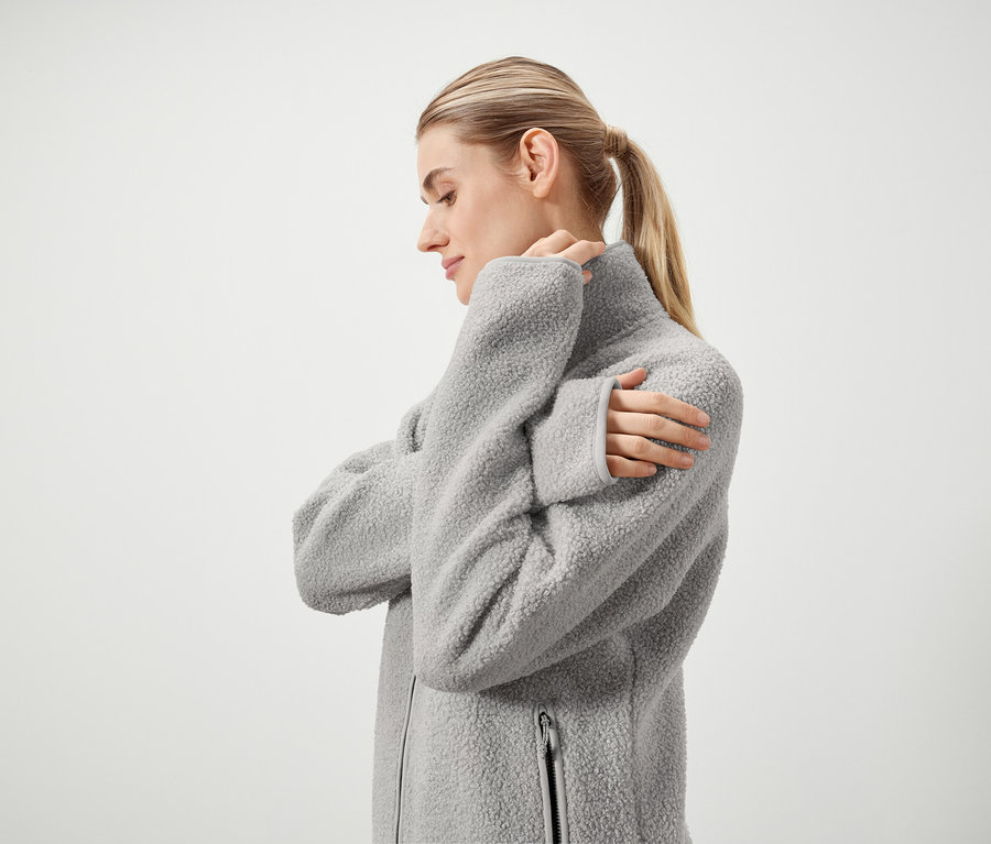 Egy szőke hajú, lófarokba kötött nő profilja, aki szürke, bolyhos, cipzáras kabátot visel, fehér háttér előtt állva.