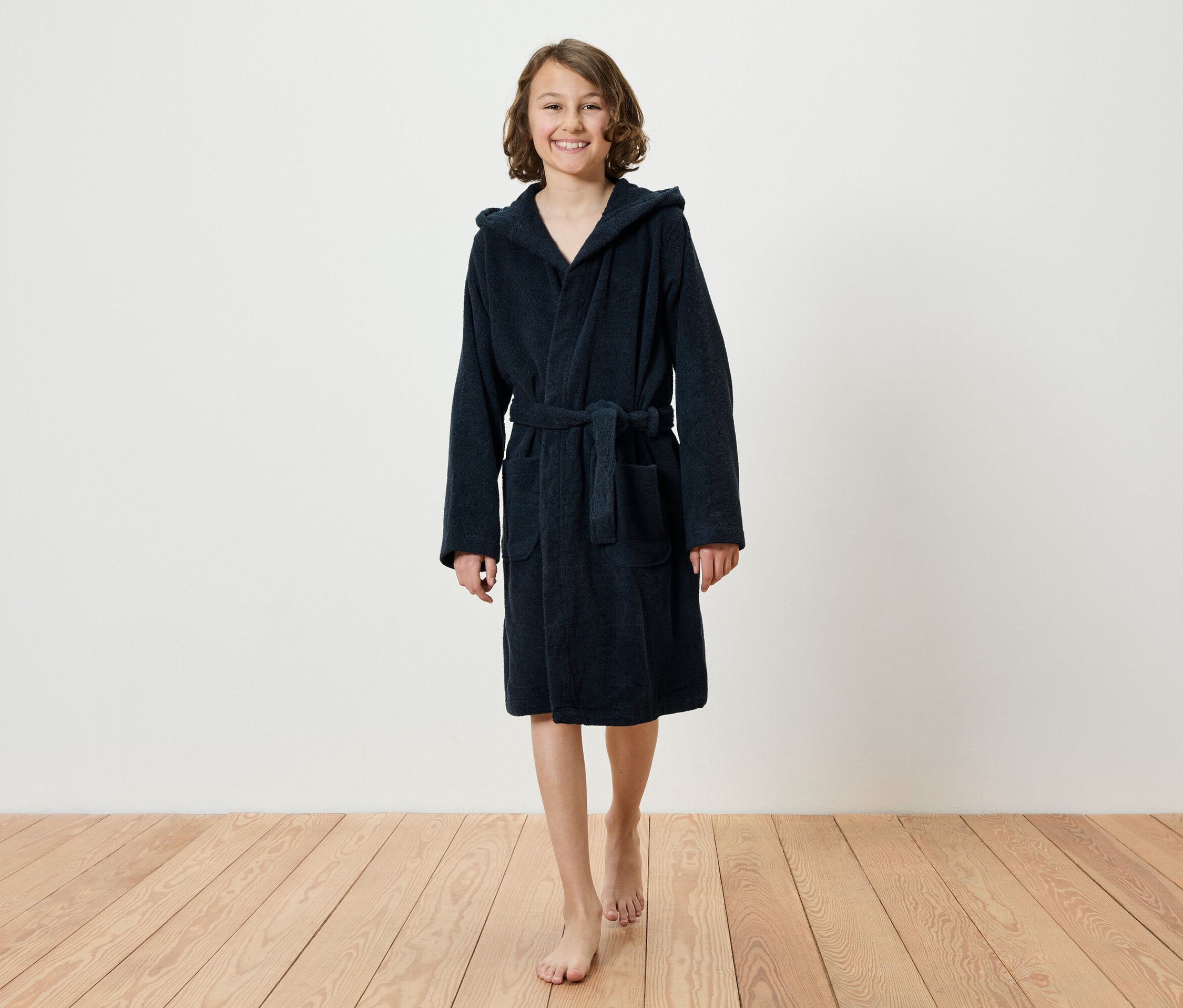 Mosolygó tinédzser lány mezítláb sétál egy fapadlón, sötétkék, kapucnis fürdőköpenyt visel.