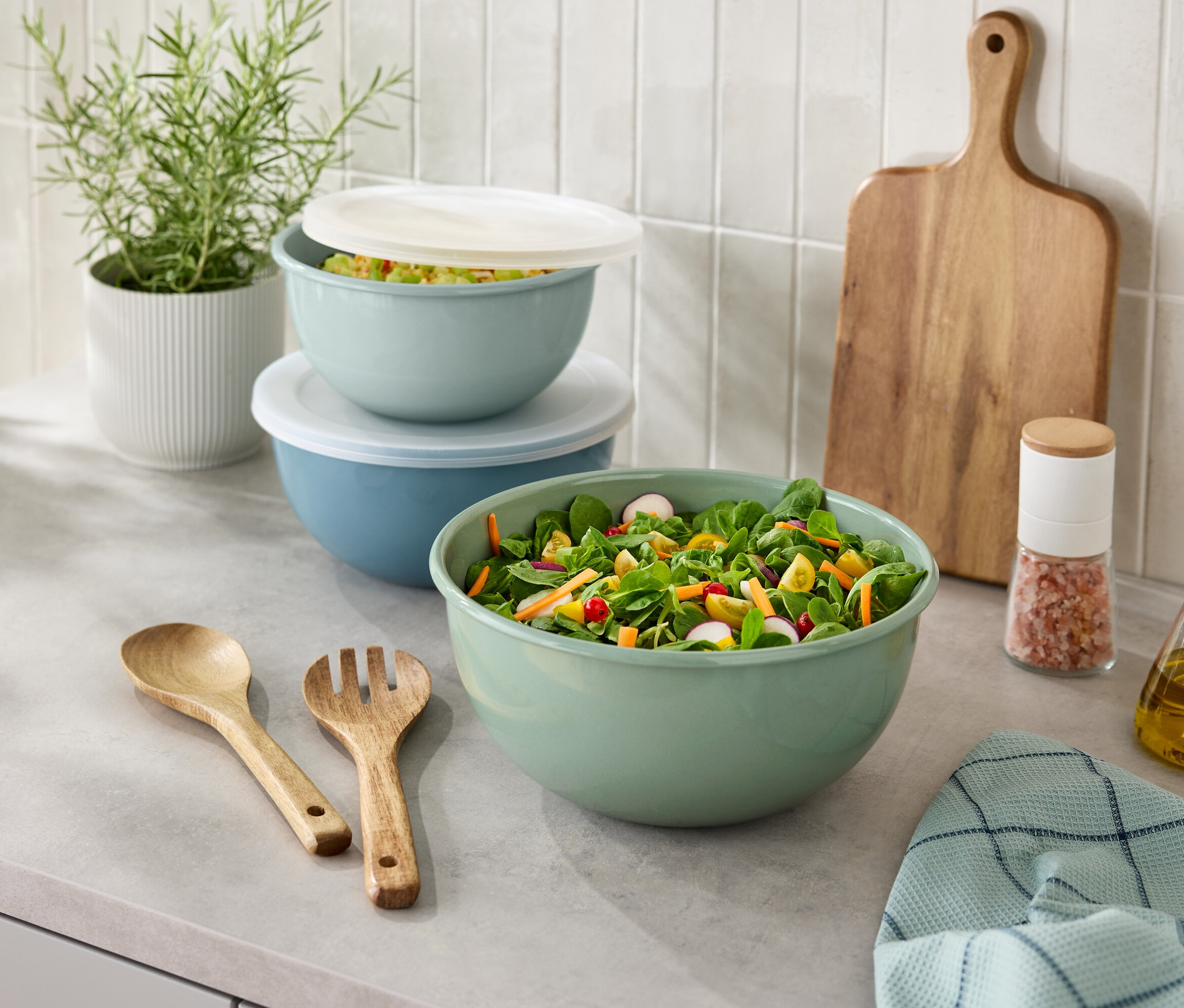 A pulton egy salátástál, fa evőeszközök, egy sótartó, egy üveg olaj, egy vágódeszka és egy cserepes növény látható.