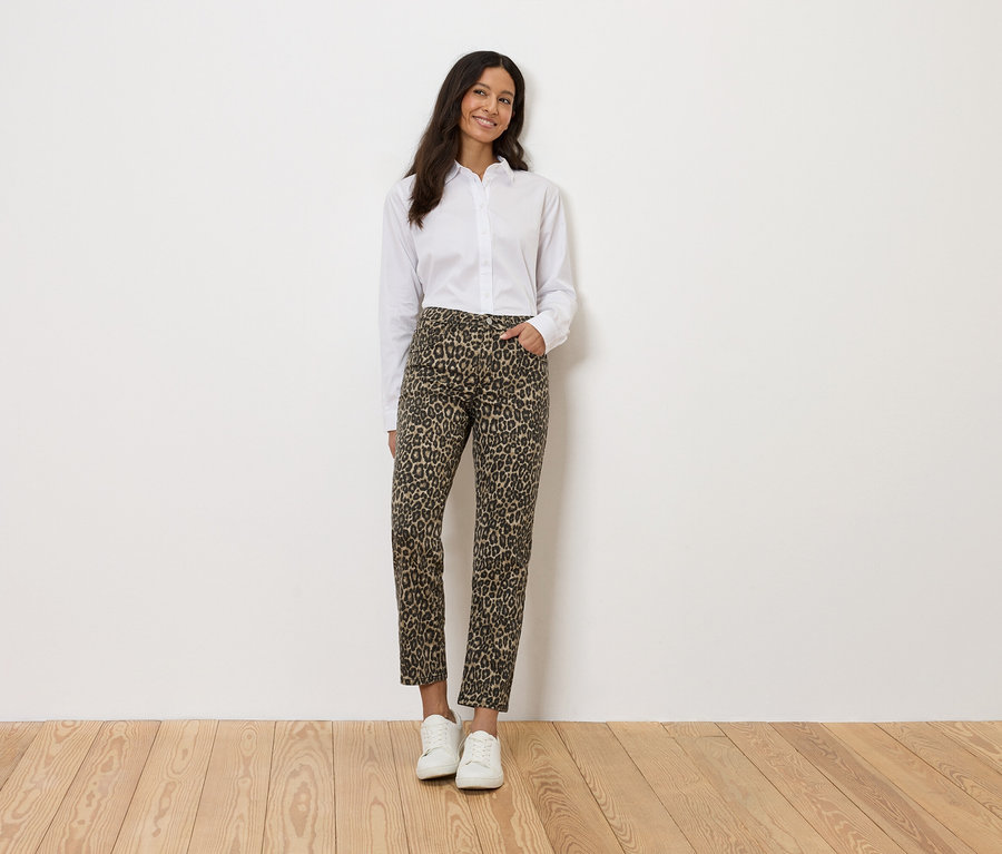 Nő fehér ingben és leopárdmintás nadrágban, fehér fal előtt állva.