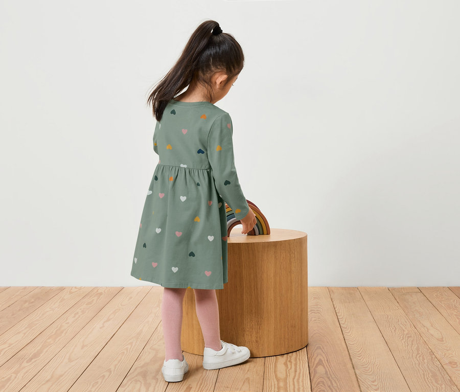 Szív mintás zöld ruhás lány áll egy fa asztalnál, amin egy szivárvány van.