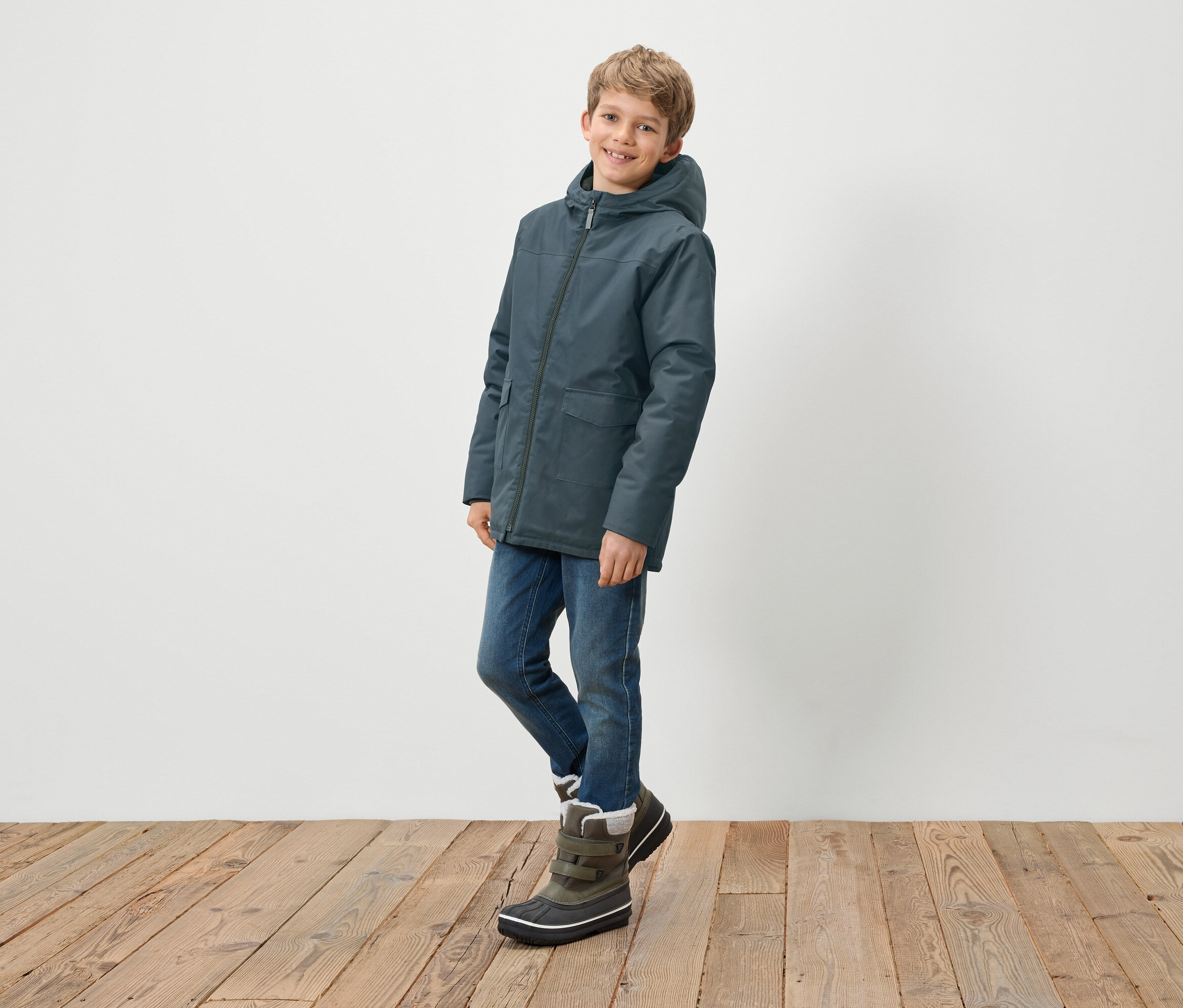 Fiú pózol kék kapucnis kabátban, farmerben és téli csizmában.