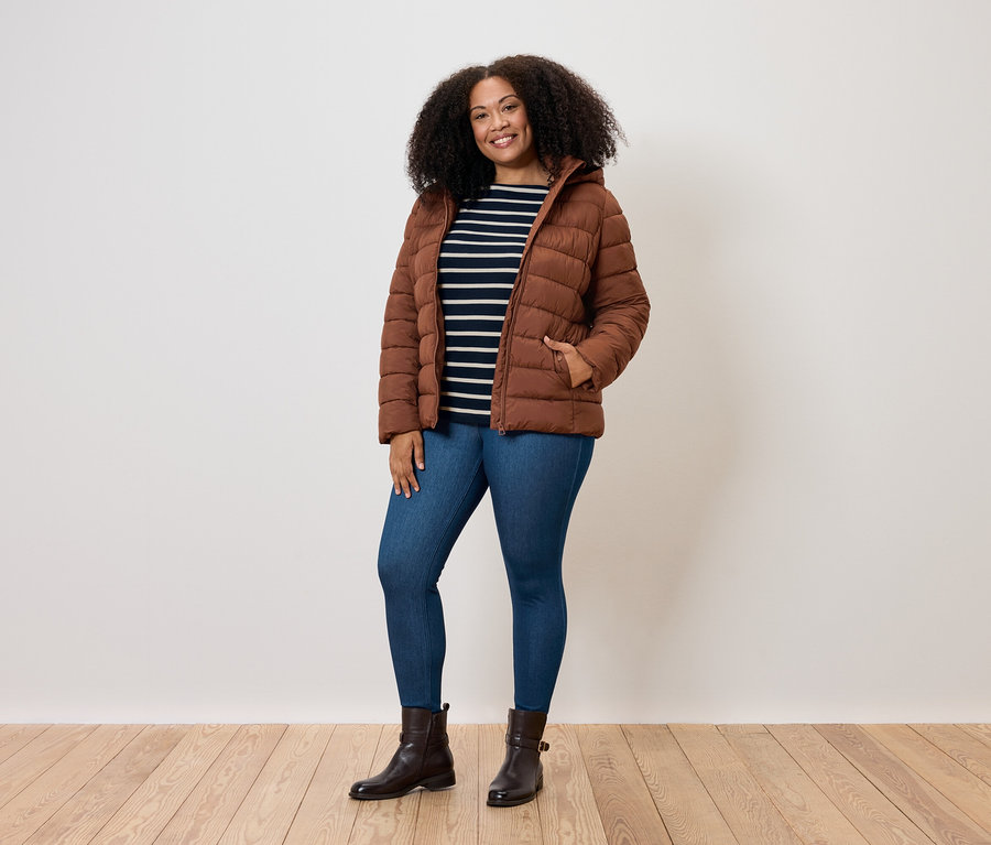 Göndör hajú nő pózol barna steppelt kabátban, kék farmerben és barna csizmában.