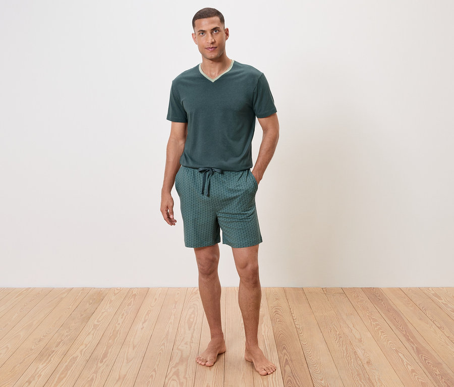Egy férfi áll mezítláb a fapadlón, zöld, rövid ujjú pizsamában és rövidnadrágban.