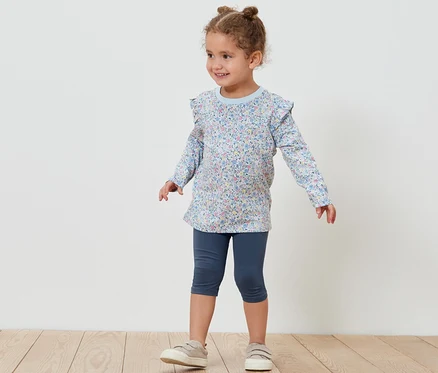 Egy kislány pózol 3/4-es leggingsben és virágos blúzban. Az 5 db 3/4-es leggings gyerekeknek egyik darabja látható.