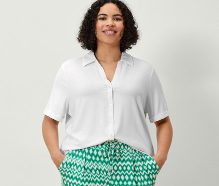 Egy nő áll, kezét a zsebében tartja, nyomott mintás 3/4-es jersey nadrágot és fehér pólóblúzt visel.