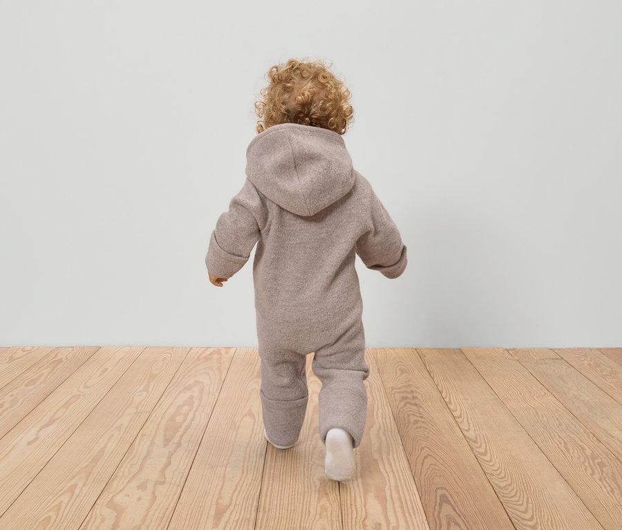 Hátulnézet egy göndör hajú kisgyermekről, aki bézs színű, kapucnis gyapjú overált és fehér zoknit visel egy fapadlón.