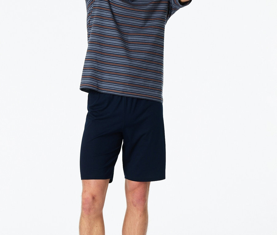 Egy férfi mezítláb áll felemelt kézzel, csíkos pólóban és kék rövidnadrágban fehér háttér előtt.