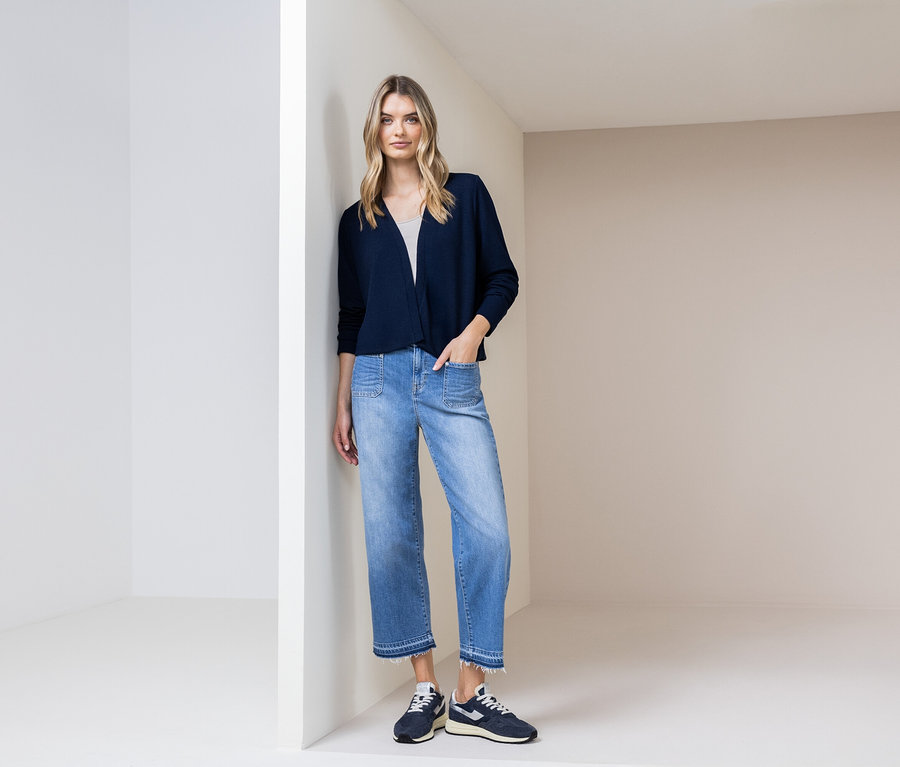 Egy nő a falnak támaszkodik, sötétkék kardigánt, kék farmert és sötétkék cipőt visel.