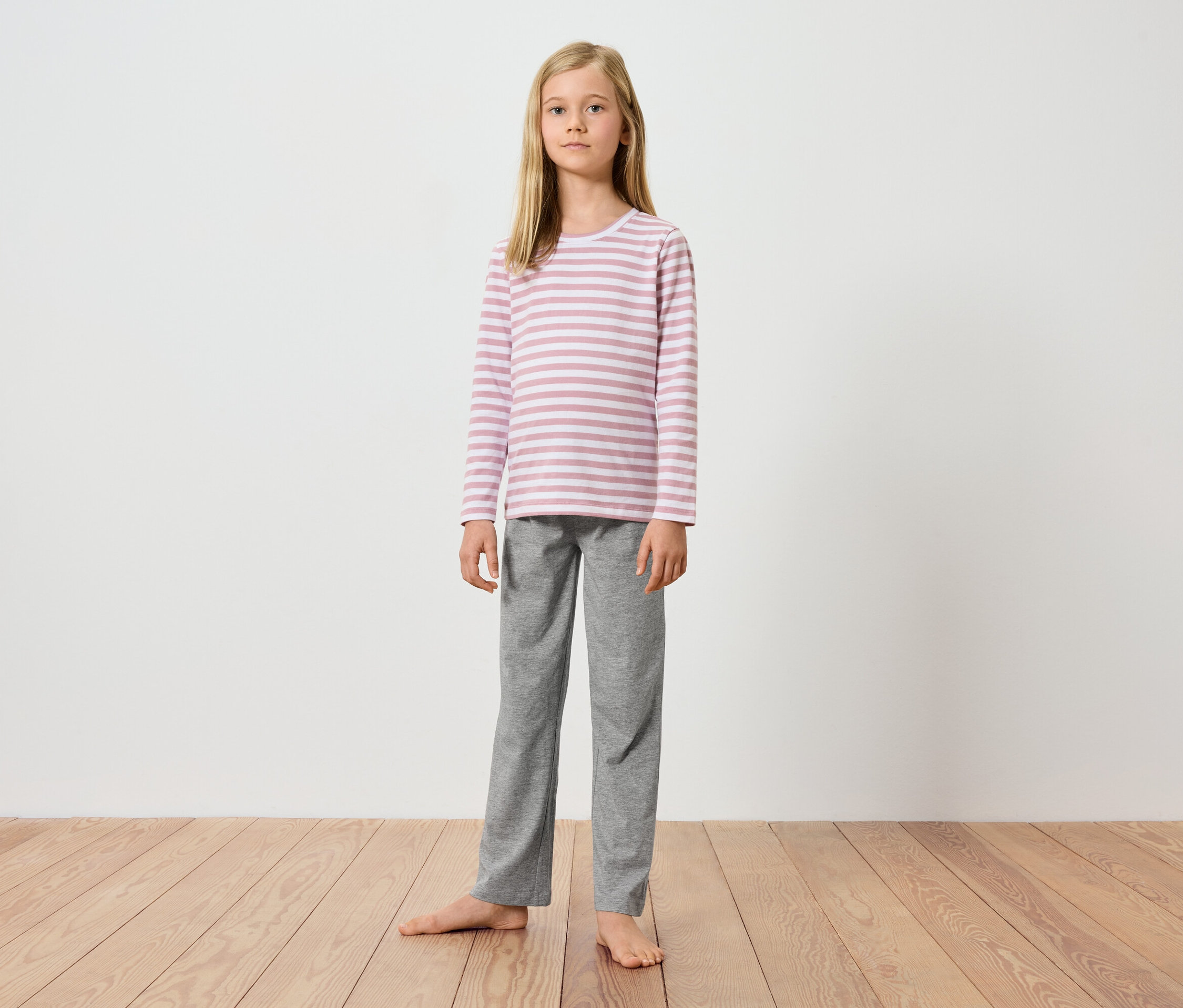 Egy fiatal lány áll mezítláb egy fapadlón egy fehér fal előtt. Rózsaszín-fehér csíkos, hosszú ujjú felsőt és szürke nadrágot visel.