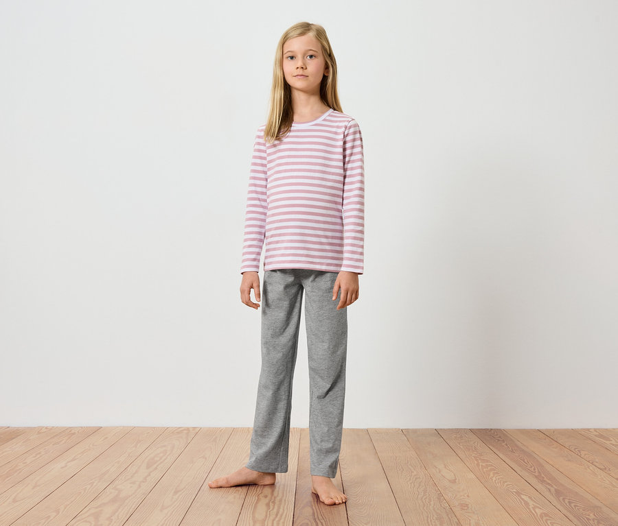 Egy fiatal lány áll mezítláb egy fapadlón egy fehér fal előtt. Rózsaszín-fehér csíkos, hosszú ujjú felsőt és szürke nadrágot visel.