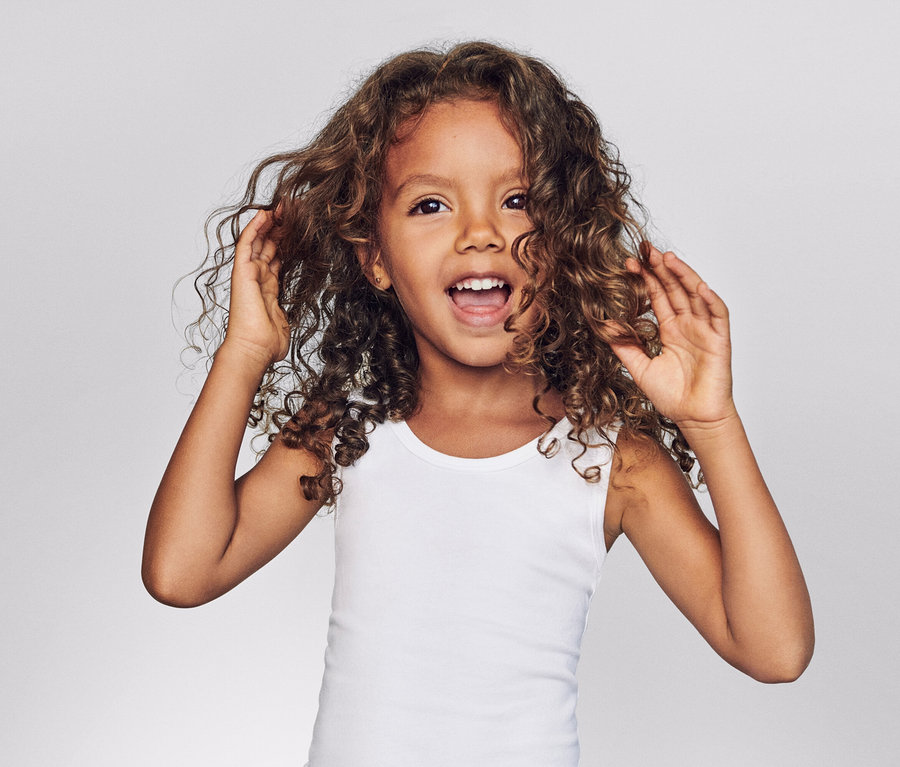 Egy göndör hajú, fehér trikós kislány nevet és megérinti a haját szürke háttér előtt.