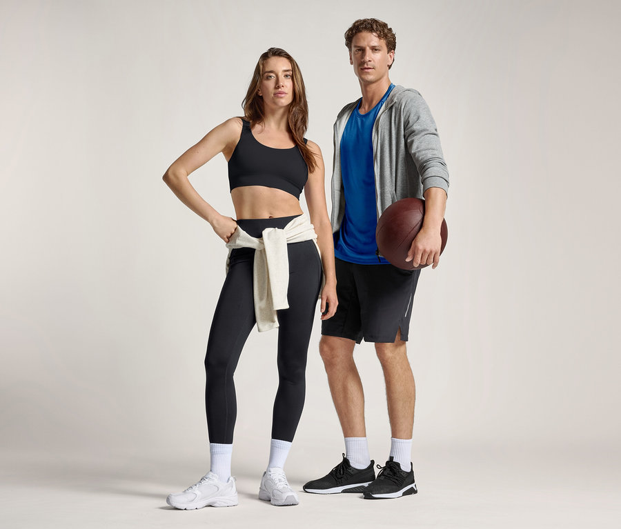 Fekete sportmelltartót és fekete női sportnadrágot viselő nő, derekára kötött pulóverrel, egy kék pólós, szürke kapucnis pulóveres és rövidnadrágos férfi mellett áll, aki amerikai futball labdát tart.