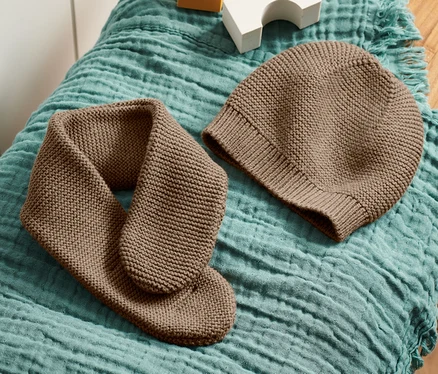 Egy barna kötött sapka és sál egy fa zsámolyon, egy kékeszöld anyagon fekszik, fehér és sárga kockák mellett.
