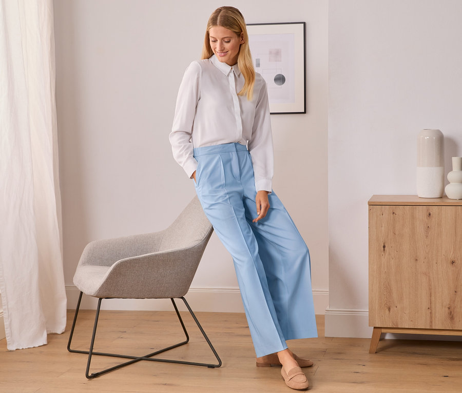 Egy nő fehér ingben és világoskék, szövött, bő szárú nadrágban dől egy szürke széknek.