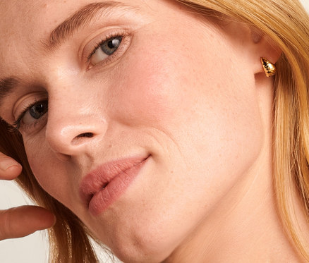 Szőke nő kezével a fején 585-ös arany Singapur láncot és 375-ös arany, kúpos, gyémántozott félkarika fülbevalót visel.