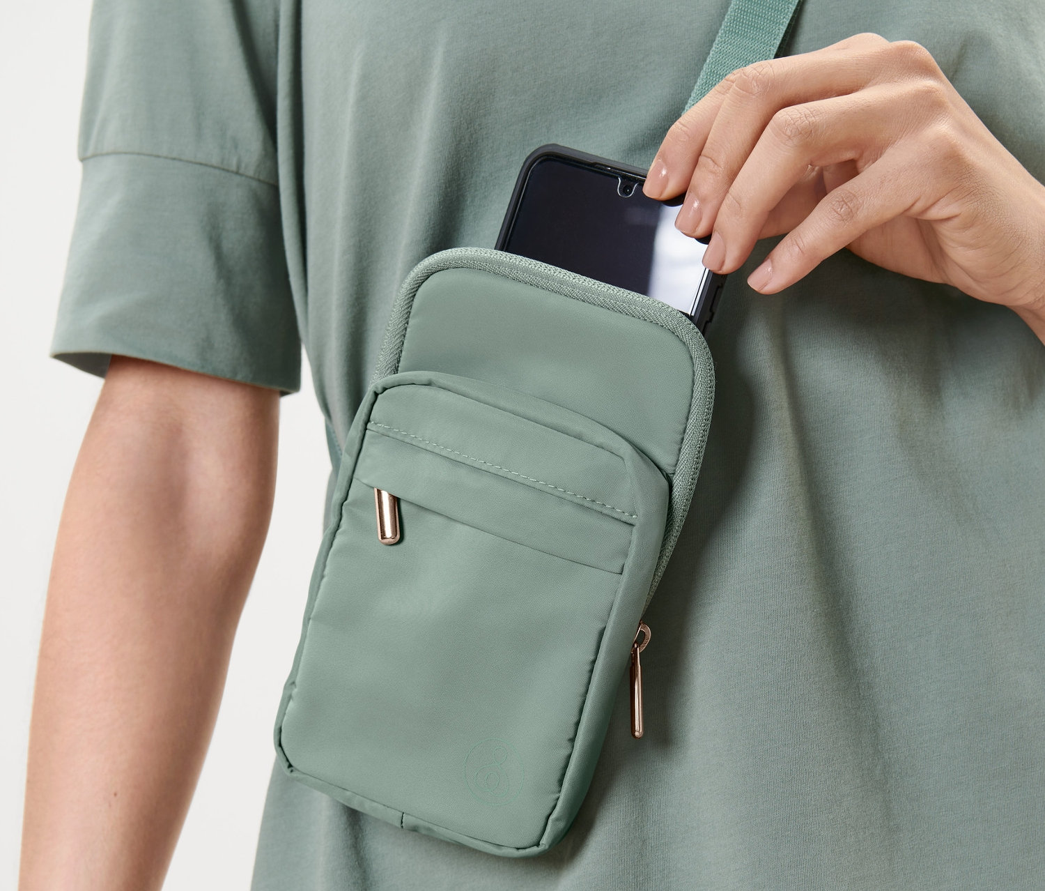 Zöld pólót viselő nő telefont tesz egy zöld oldaltáskába.
