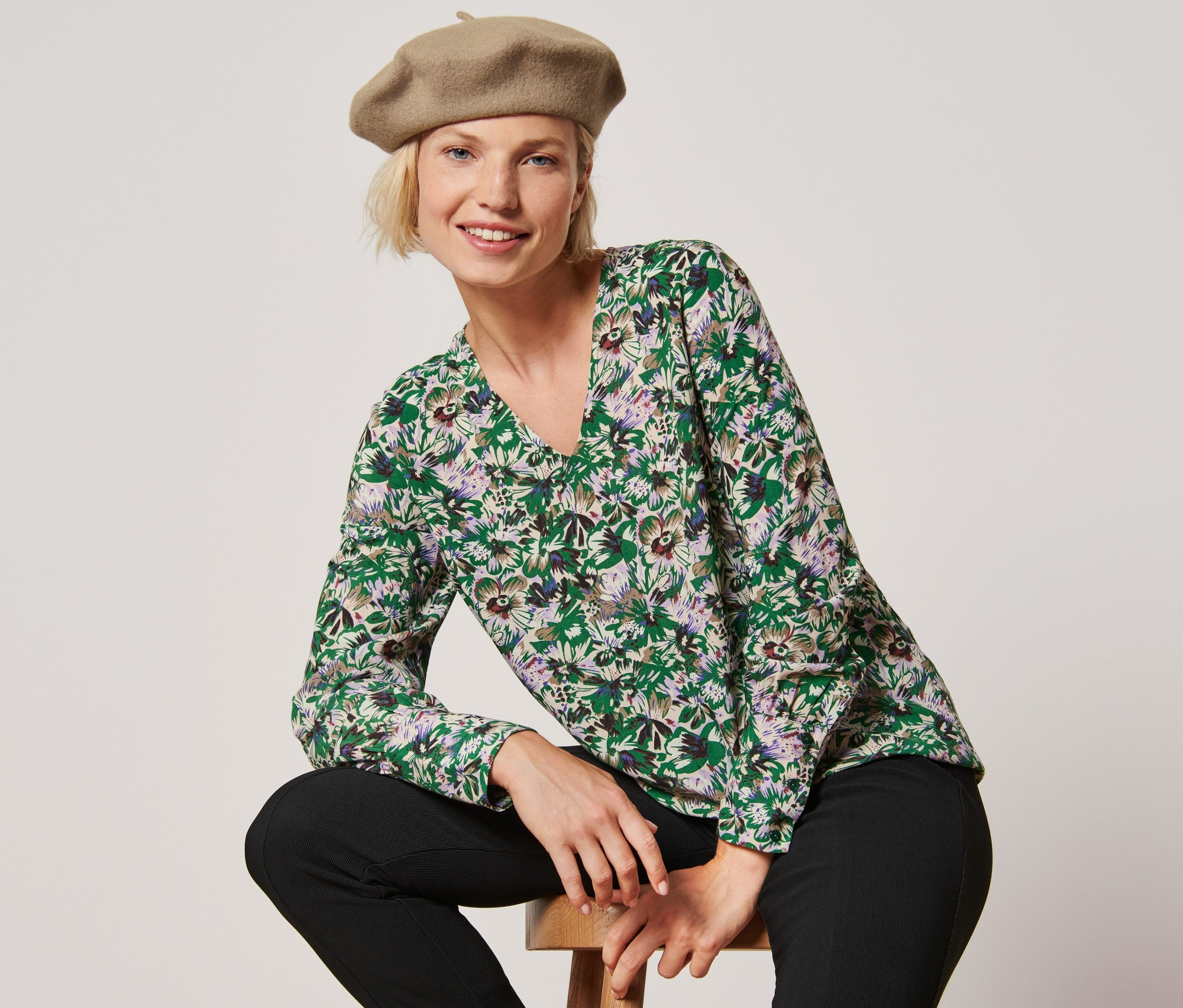 Egy nő egy széken ül, virágos női blúzt és egy sapkát visel.