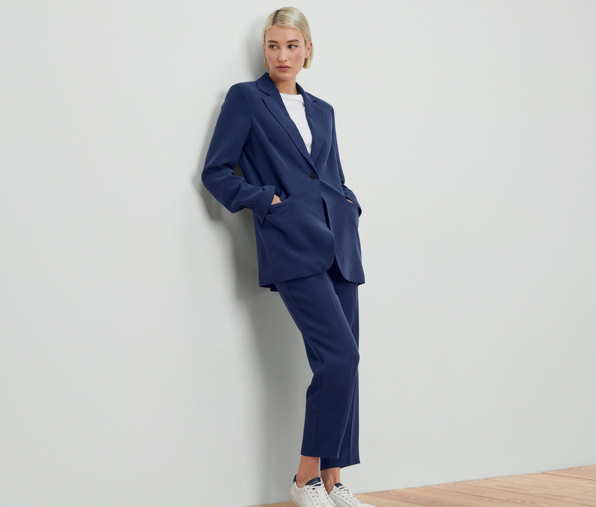 A falnak támaszkodó nő kék szövött blézert és hozzá illő nadrágot visel.