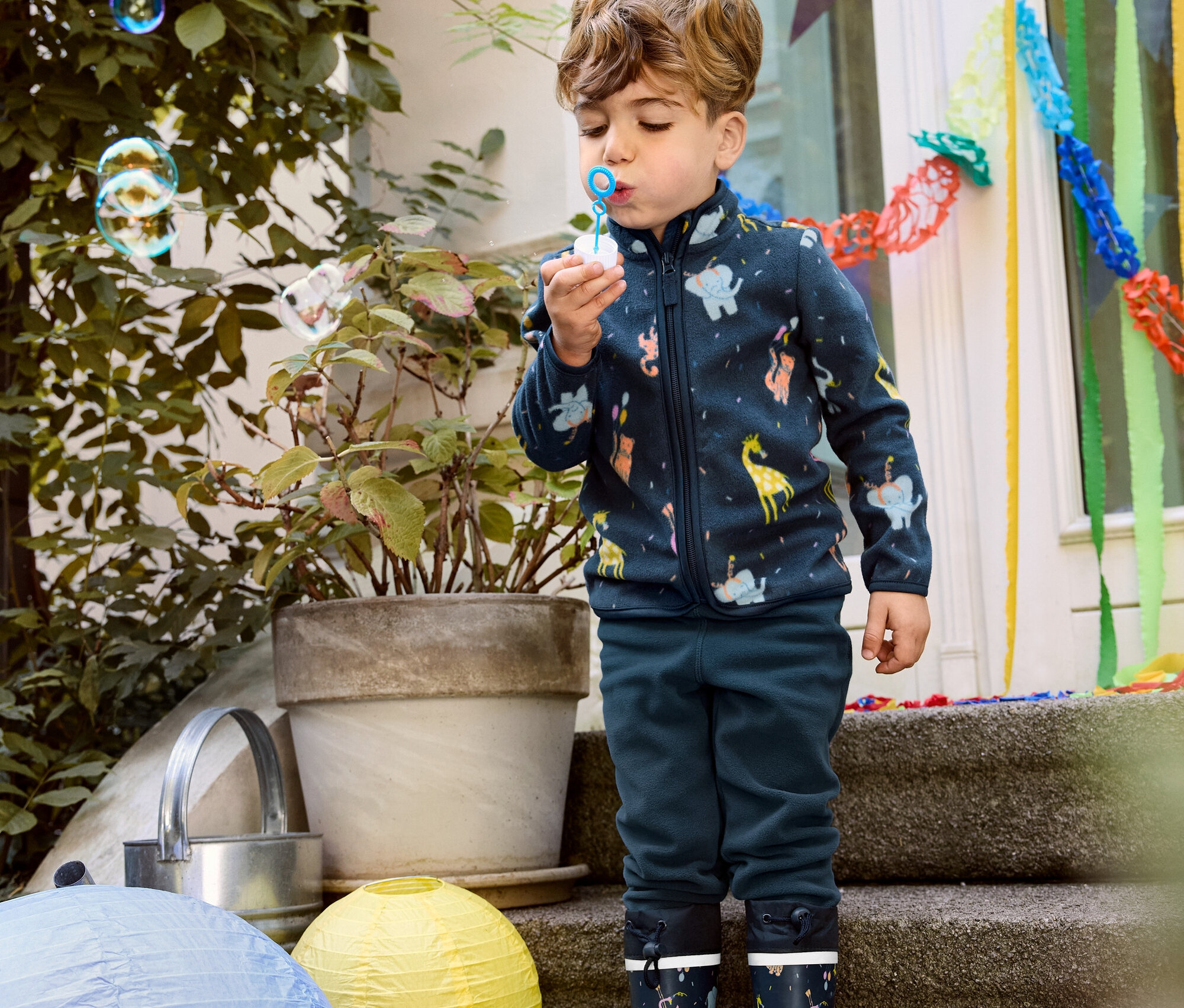 Fiú buborékot fúj, állatos kisgyerek polárfelsőt, sötétkék polárnadrágot és gumicsizmát visel.