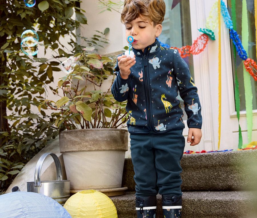 Fiú buborékot fúj, állatos kisgyerek polárfelsőt, sötétkék polárnadrágot és gumicsizmát visel.