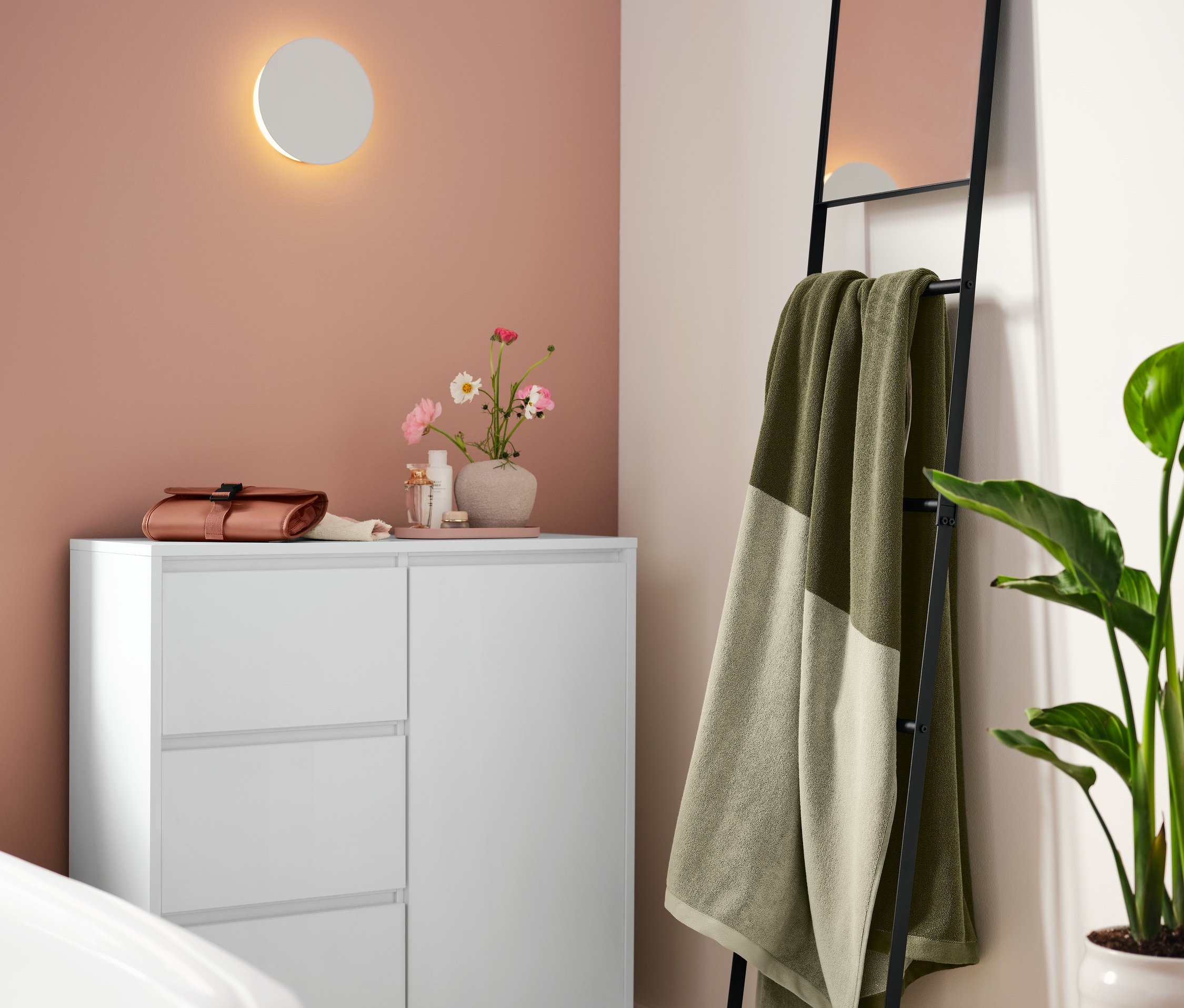 A fürdőszobában egy fehér fürdőszobai komód áll, rajta egy rozsdaszínű fém tálca és egy neszesszer. A falon egy LED-es fali lámpa akkumulátorral. A fal mellett egy törölközőtartó létra tükörrel, rajta egy XL-es zöld fürdőlepedő.