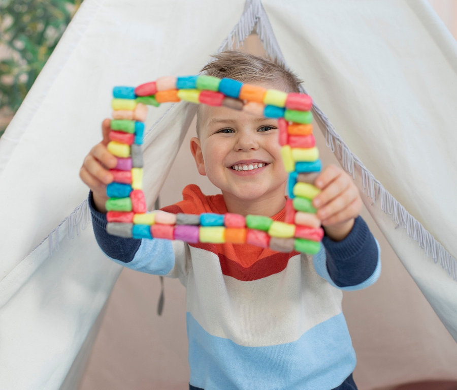 Egy fiú ül egy teepee-ben, és egy színes négyzetet tart a kezében a PlayMais®-Setből.