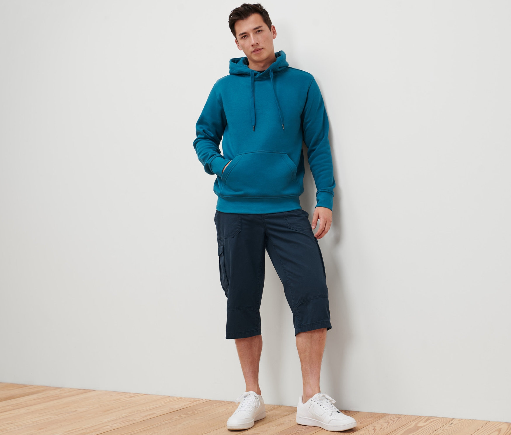 Férfi támaszkodik a falnak, kékeszöld kapucnis pulóvert és sötétkék férfi 3/4-es bermudanadrágot visel.