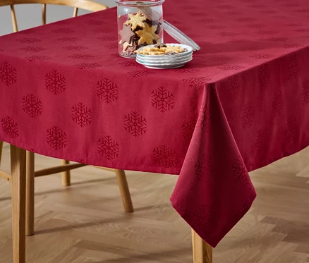 Egy piros, hópehely mintás jacquard asztalterítő látható az asztalon, mellette egy üveg sütemény és egy tányér édesség.