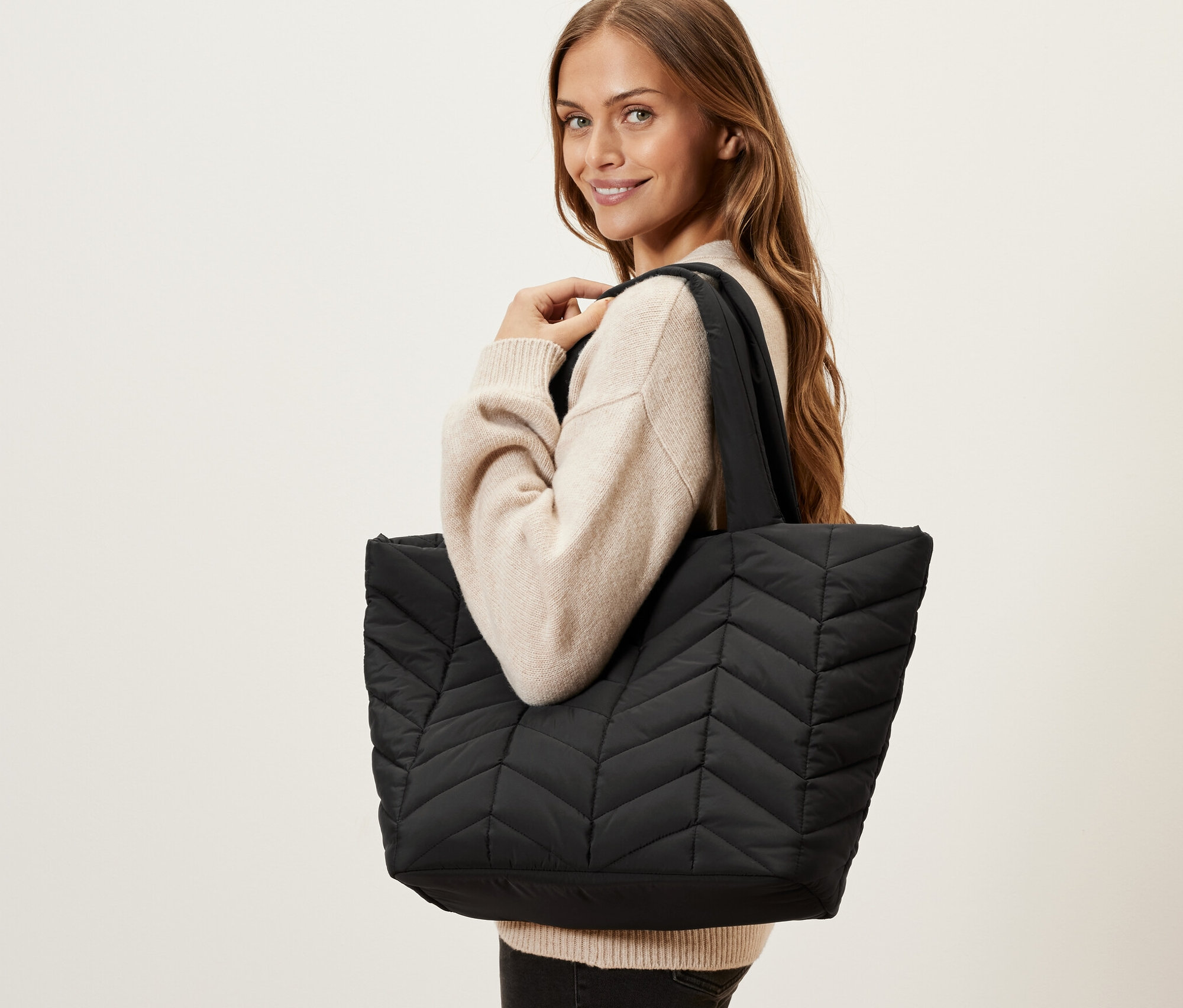 Nő pózol fekete, steppelt shopper táskával a vállán.