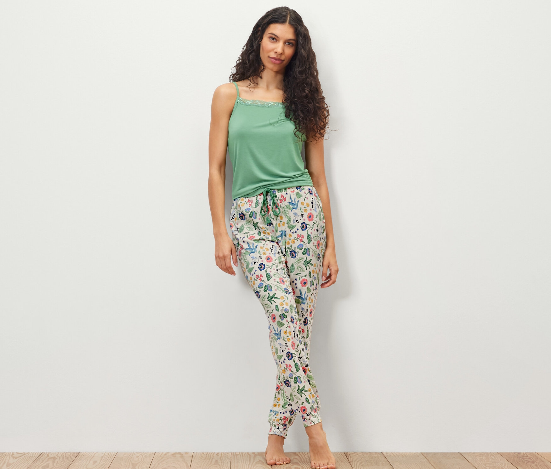 Egy nő áll egy fehér fal előtt, zöld felsőt és virágos női pizsamanadrágot visel.