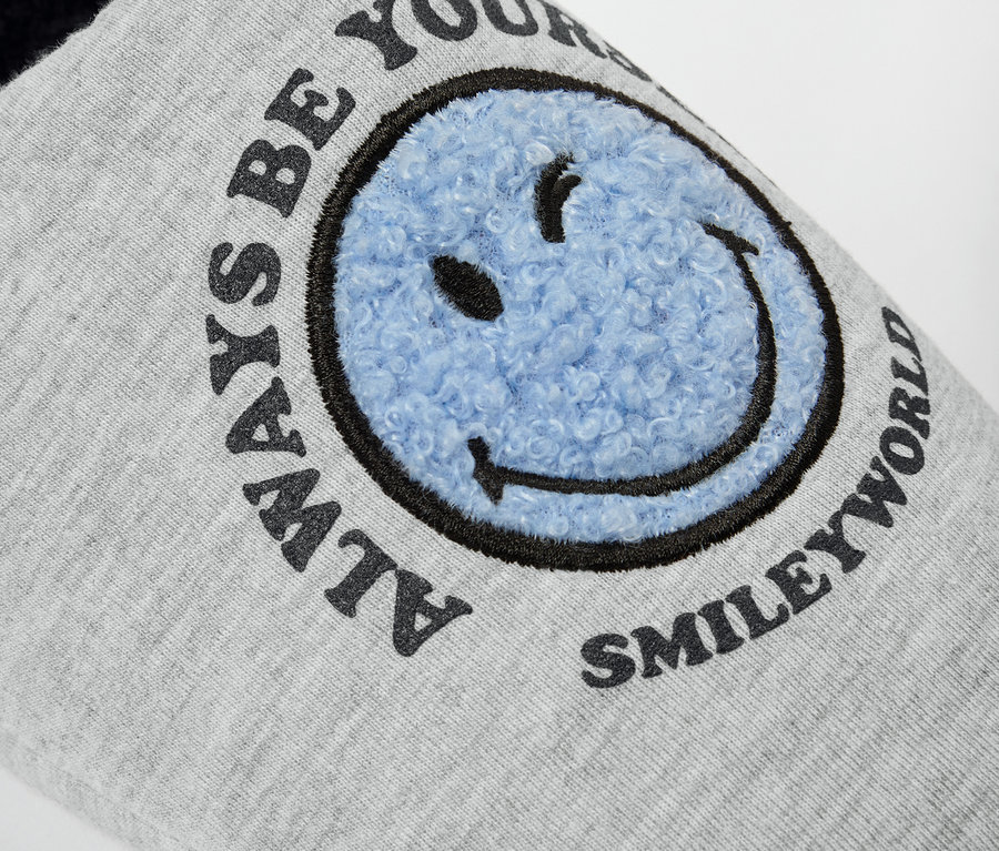 Egy szürke SMILEYWORLD® házicipő részlete, kék mosolygós arccal.