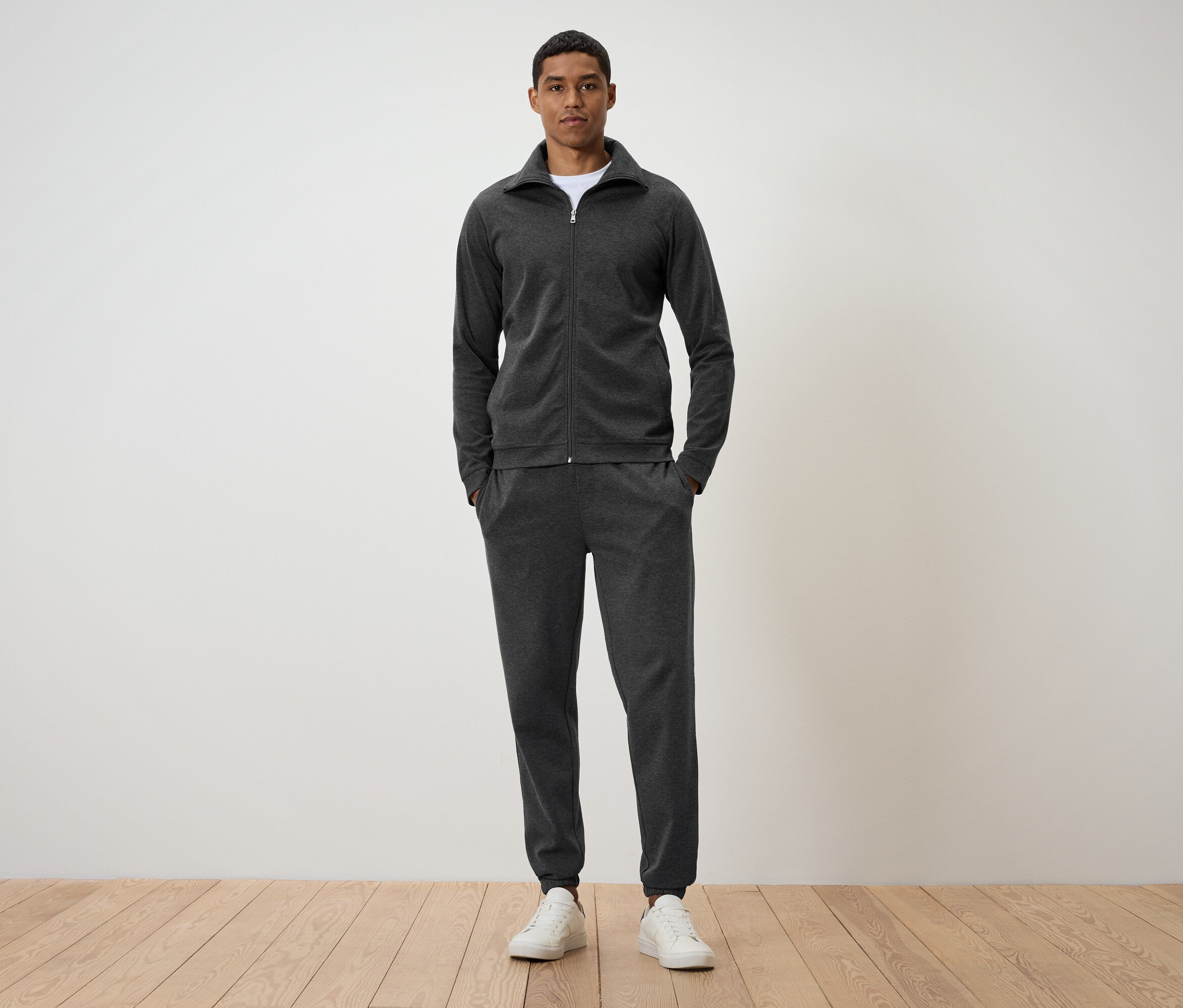 Egy férfi áll egy fapadlón, sötétszürke melegítőben és fehér tornacipőben.