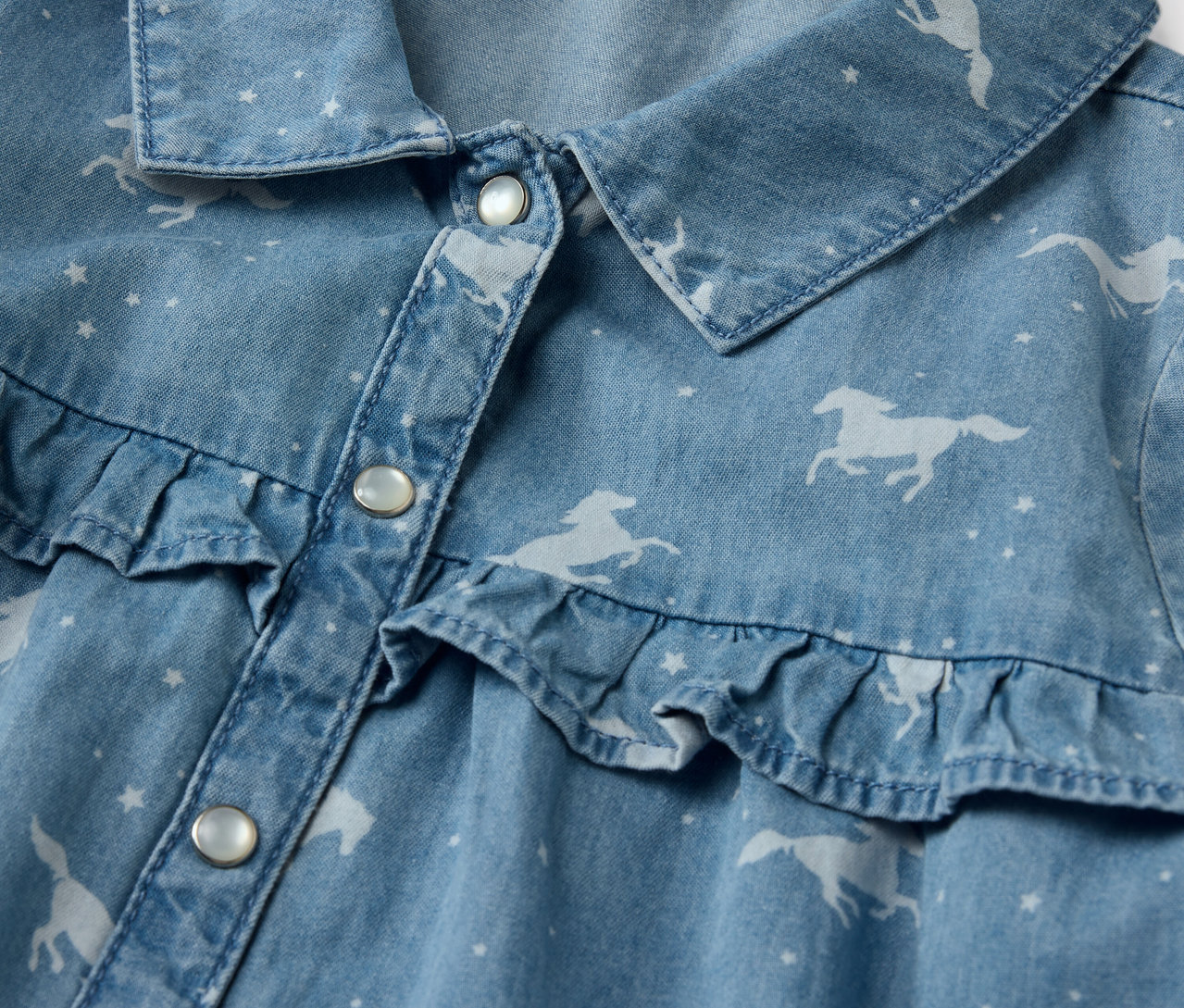 Egy gyermek farmerruhájának részlete, lovas mintával és fodrokkal.