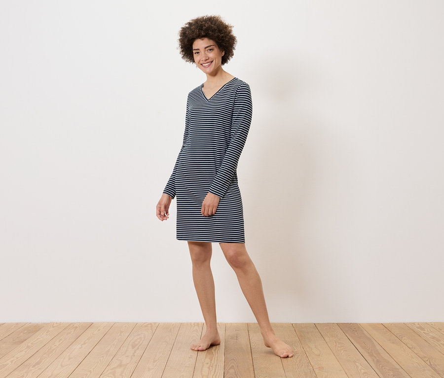 Egy nő áll mezítláb a fapadlón, és egy csíkos, hosszú ujjú ruhát visel.