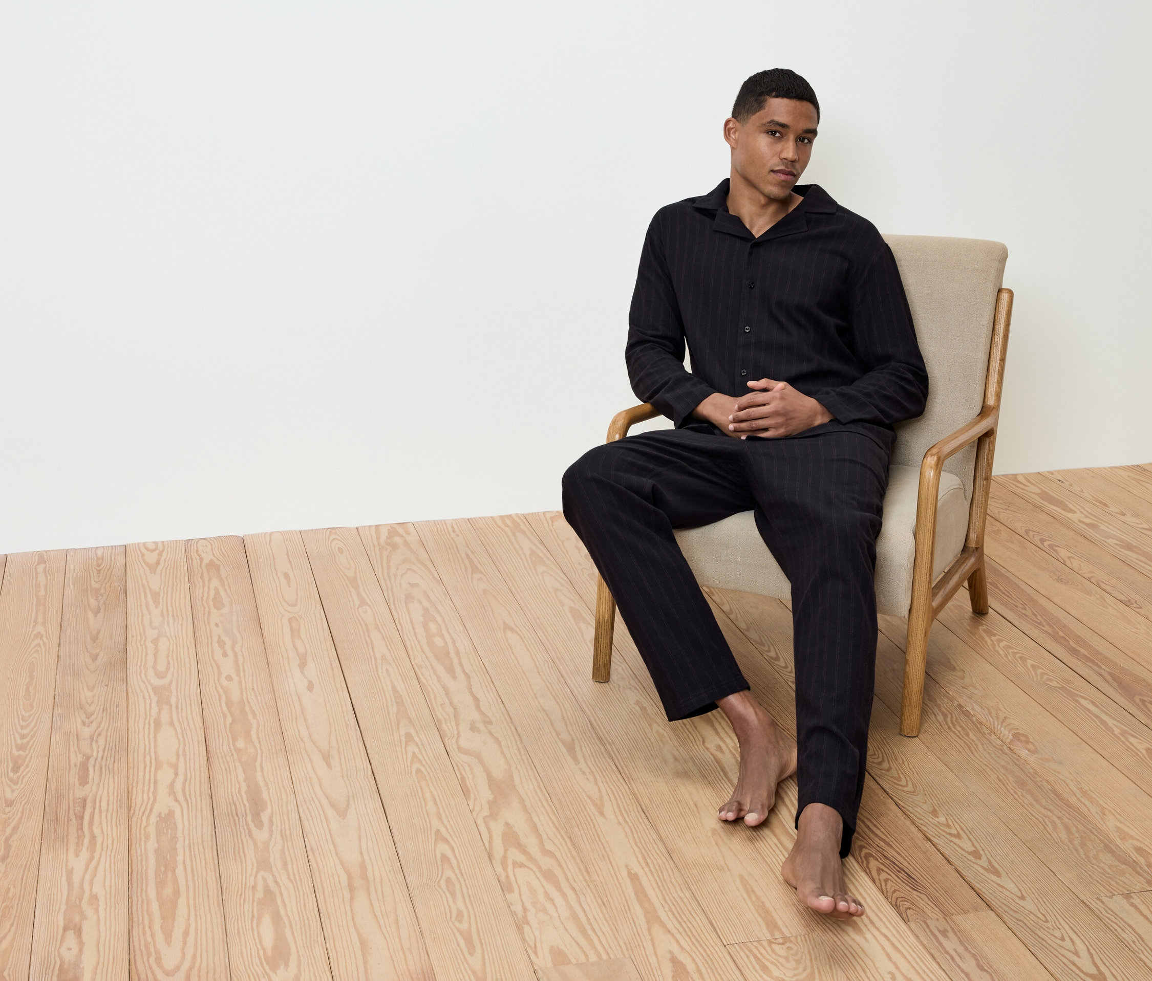 Férfi ül mezítláb egy bézs fotelben a fapadlón, fekete csíkos pizsamában.