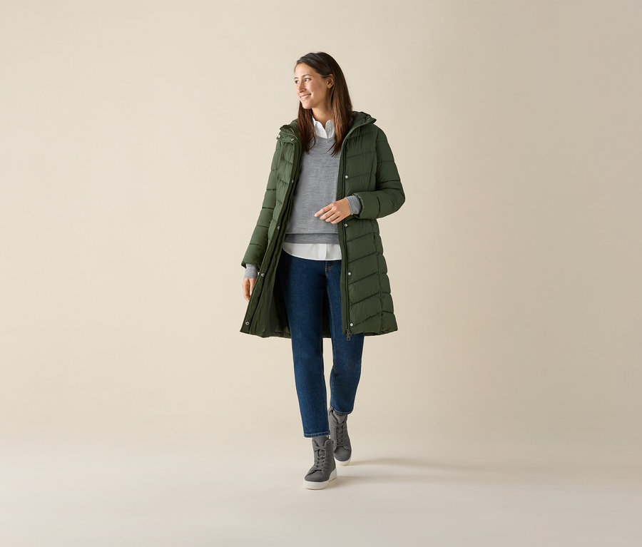 Nő visel szürke finom kötésű merinó pulóvert, zöld kabátot és farmert.