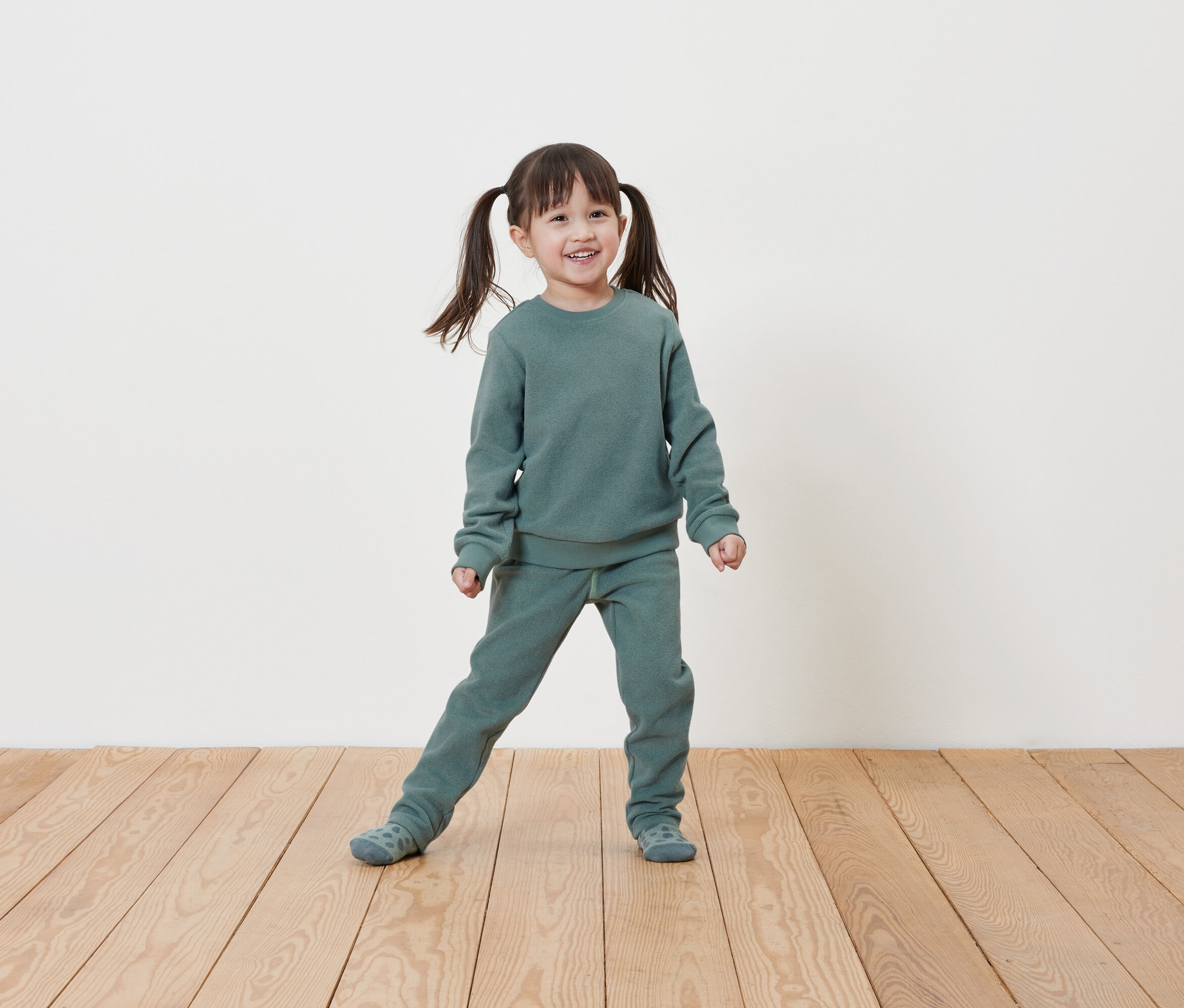 Mosolygó kislány copfokkal világoszöld pulóverben és jogging nadrágban pózol a fapadlón egy fehér fal előtt.