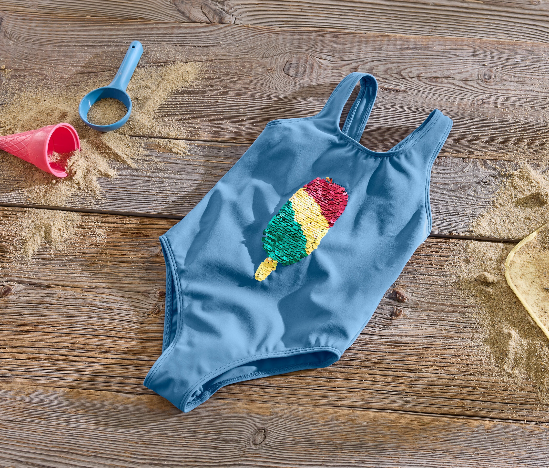 Kék gyerek fürdőruha átfordítható flitterekkel fa felületen, homokkal és játékokkal.