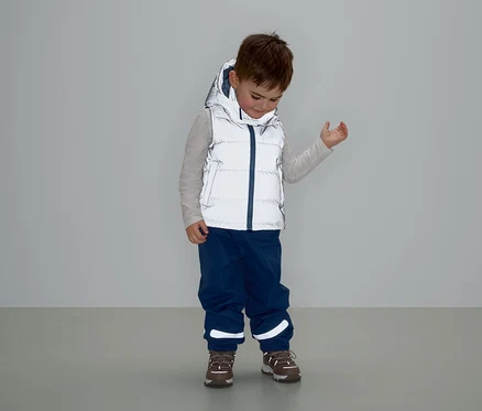 Egy fiú szürke, kapucnis mellényben, kék nadrágban és barna cipőben áll és lefelé néz.