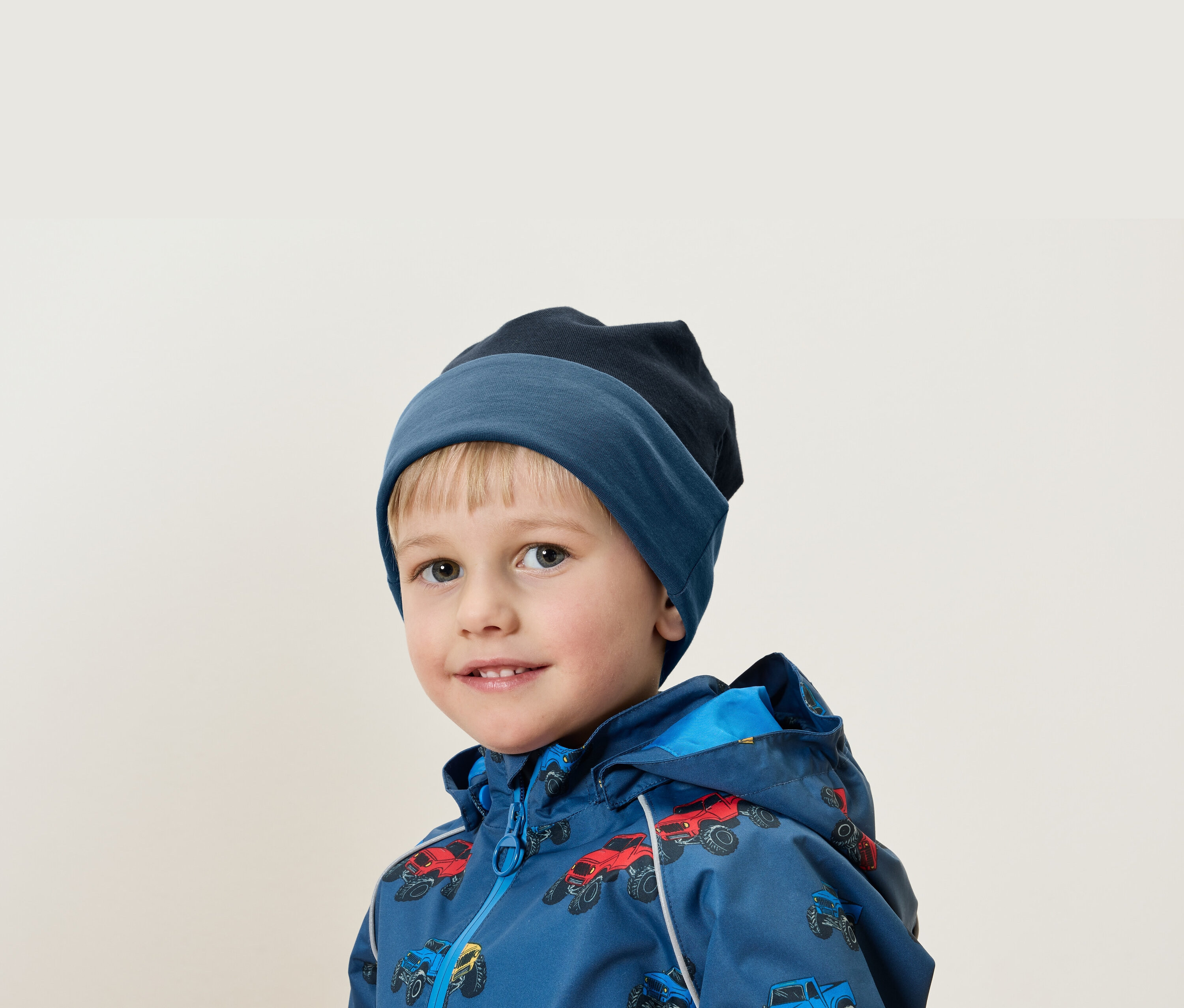 Portré egy szőke hajú fiúról kék sapkában és autós mintás kabátban.
